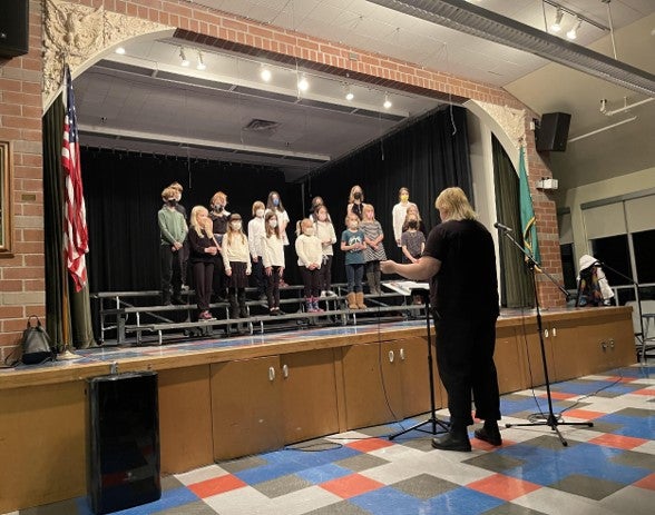 student choir practicing on stage with teacher instructing below